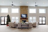 This generously sized single-floor space is designed as an open, multi-use common area that balances comfort with functionality. The centerpiece of the main lounge is a dramatic floor-to-ceiling stacked stone fireplace fitted with a mounted flat-screen television and a rustic wood mantel, creating a warm and inviting focal point. Surrounding the fireplace, neutral-toned upholstered seating is arranged around a farmhouse-style coffee table, encouraging relaxed social interaction.

The soaring ceilings, lined with a row of clerestory windows along multiple walls, allow an abundance of natural light to fill the interior throughout the day. Dual ceiling fans circulate air efficiently across the expansive open floor plan, which is finished with neutral patterned carpet throughout. Exposed ductwork along the upper walls adds an industrial accent that contrasts with the otherwise warm, contemporary aesthetic.

Multiple exterior entry points, including glass-paneled double doors, connect the space to the outdoors. A quieter activity alcove features light wood tables and chairs, well-suited for games, puzzles, or small group gatherings. Additional open areas near entry corridors provide flexible space for various uses. The overall ambiance is bright, welcoming, and thoughtfully designed for communal living.