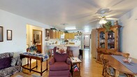 This single-floor home exudes a warm, lived-in charm with a thoughtfully arranged open-concept layout that seamlessly connects the main living and kitchen areas. The kitchen is well-appointed with honey-toned wood cabinetry, a white appliance suite including a refrigerator and microwave, and a peninsula with bar stool seating that serves as a natural dividing point between cooking and living spaces. A large overhead fluorescent fixture provides ample task lighting over the kitchen workspace, complementing the natural warmth of the wood tones throughout. The living area features light hardwood flooring that runs throughout, lending cohesion and a sense of spaciousness to the open floor plan. A ceiling fan with a light kit keeps the space comfortable and well-lit. Classic wood furniture, including an ornate display cabinet and traditional dining pieces, add a homey, country-inspired aesthetic. A hallway visible from the main living area leads to additional rooms, and a wood-paneled entry door provides a welcoming focal point at the end of the corridor. The overall ambiance is cozy and functional, with a layout that encourages easy flow between daily living spaces.