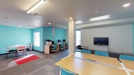 This single-floor study room is thoughtfully designed to support both individual and group learning. The space is defined by its bright, energizing teal accent walls that contrast cleanly against neutral white and gray tones, creating an atmosphere that is both stimulating and focused. Recessed and linear ceiling lighting ensures consistent, even illumination throughout the room, complemented by natural light from windows along the perimeter walls.

The room is organized into distinct functional zones. A dedicated computer workstation area lines one wall, featuring a row of desktop computers set on light wood desks with matching teal task chairs, alongside a multifunction printer, making it a fully equipped resource hub. Opposite this area, a large flat-screen television mounted on the wall serves as a presentation or collaboration display.

At the center and foreground of the space, long communal tables paired with teal chairs provide ample seating for group study or individual work. The carpeted flooring, featuring a neutral grid pattern with a red accent rug in one corner, adds a layer of comfort and warmth underfoot. Structural columns are integrated seamlessly into the layout, and a storage credenza near the display screen offers additional organizational capacity. Overall, the space is clean, modern, and highly functional.