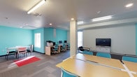 This single-floor study room is thoughtfully designed to support both individual and group learning. The space is defined by its bright, energizing teal accent walls that contrast cleanly against neutral white and gray tones, creating an atmosphere that is both stimulating and focused. Recessed and linear ceiling lighting ensures consistent, even illumination throughout the room, complemented by natural light from windows along the perimeter walls.

The room is organized into distinct functional zones. A dedicated computer workstation area lines one wall, featuring a row of desktop computers set on light wood desks with matching teal task chairs, alongside a multifunction printer, making it a fully equipped resource hub. Opposite this area, a large flat-screen television mounted on the wall serves as a presentation or collaboration display.

At the center and foreground of the space, long communal tables paired with teal chairs provide ample seating for group study or individual work. The carpeted flooring, featuring a neutral grid pattern with a red accent rug in one corner, adds a layer of comfort and warmth underfoot. Structural columns are integrated seamlessly into the layout, and a storage credenza near the display screen offers additional organizational capacity. Overall, the space is clean, modern, and highly functional.