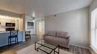 This single-floor residence offers a thoughtfully designed layout that connects the main living spaces in a functional, open arrangement. The living room serves as the central gathering area, featuring wood-look flooring, a textured ceiling, a ceiling fan, and a tile-surround fireplace flanked by two windows that allow natural light to filter in. A glass-top coffee table and neutral-toned sofa complement the clean, understated aesthetic throughout.

The kitchen is positioned adjacent to a breakfast bar that provides casual seating and a natural divide from the dining area. It is equipped with gray cabinetry, white countertops, a subway tile backsplash, black appliances including a range, microwave, and refrigerator, as well as a stainless steel dishwasher. A compact dining space with a round table and chairs sits nearby, conveniently located beside an in-unit laundry closet fitted with a full-size washer and dryer topped by additional gray cabinetry for storage.

The bedroom receives natural light through a large sliding glass door with vertical blinds and includes a ceiling fan overhead. The bathroom features a tub-shower combination with white tile surround, wood-look flooring, and clean, minimal fixtures. A small entryway with a bench and security panel completes the layout.