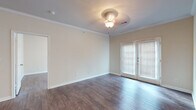 This single-floor space is thoughtfully laid out and finished with consistent dark hardwood-style flooring throughout, lending a cohesive, warm aesthetic across all rooms. The main living area is a standout feature, showcasing elegant crown molding, neutral cream-toned walls, and a decorative ceiling light fixture. French doors flood the space with natural light and provide access to the exterior, enhancing the open, airy feel of the room.

Additional rooms offer ample space, each finished with the same hardwood-style flooring and clean white baseboards. Ceiling fans with light fixtures are present in select rooms, adding both functionality and a classic touch. Windows fitted with blinds allow for adjustable natural light throughout.

The kitchen is well-equipped, featuring rich warm-toned wood cabinetry, granite-style countertops, a full-size refrigerator, and a double pantry or storage closet. The galley-style layout maximizes counter and cabinet space efficiently.

The bathroom includes a full soaking tub with a tiled surround, a white vanity with storage, and direct access to a walk-in closet, adding practical convenience. Overall, the space presents a clean, move-in-ready environment with classic detailing.