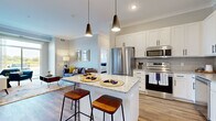 This thoughtfully designed 3-bedroom, 2-bathroom residence offers a functional and stylish single-floor layout. The open-concept kitchen and living area serves as the heart of the home, anchored by a granite-topped island with bar seating and sleek pendant lighting overhead. The kitchen is outfitted with white shaker cabinetry, subway tile backsplash, stainless steel appliances — including a French door refrigerator, range, microwave, and dishwasher — and ample counter and storage space. Warm hardwood-style flooring flows throughout the main living areas, complementing the light, neutral color palette. The living room is bright and inviting, featuring large sliding glass doors that flood the space with natural light and provide outdoor views. The primary bedroom is a comfortable retreat, carpeted for warmth, with a generously sized window overlooking greenery. A dedicated walk-in closet with wire shelving provides excellent storage. The bathrooms feature granite vanities, white cabinetry with matte black hardware, hardwood-style flooring, and a glass-enclosed walk-in shower. Crown molding adds a refined touch throughout, while the overall layout efficiently balances shared living spaces with private bedroom quarters.