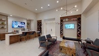 This single-floor amenity space is thoughtfully designed to offer a variety of social and recreational areas within a cohesive, contemporary aesthetic. The main lounge area features soaring ceilings accented by a striking brass sputnik chandelier, which serves as a focal point above a curated seating arrangement of mid-century modern armchairs and a distinctive live-edge wood coffee table set atop a traditional patterned area rug. Recessed lighting throughout complements the warm-toned, cream-colored walls and large-format tile flooring.

A built-in walnut shelving unit with diagonal book storage flanks a wall-mounted flat-screen television, adding both function and visual interest. Adjacent to the lounge, a bar counter with bar-height seating and wood cabinetry creates an ideal gathering spot. A separate kitchenette area provides stainless steel appliances, including a microwave and under-counter refrigerator, white tile backsplash, and open shelving for added convenience.

Accessible through a sliding barn door, a dedicated game room houses a pool table beneath elegant pendant lighting, alongside a dining table and chairs set on a patterned rug. A glass-partitioned area nearby adds an additional layer of versatility to this well-appointed communal space.