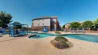 Located at 2702 N Judge Ely Blvd in Abilene, Texas, this outdoor amenity area centers around a generously sized freeform swimming pool with a distinctive figure-eight layout, offering both a main pool and an adjoining hot tub/spa. The pool deck is finished in smooth concrete and outfitted with an array of blue lounge chairs and canopied pergola structures, providing shaded relaxation spots directly alongside the water. In-pool platforms and steps make for easy entry and a resort-style experience under the wide Texas sky.

Bordering the pool area is a well-maintained clubhouse building featuring a charming pitched roof, lap siding, and a series of large grid-pane windows paired with bold red French doors that open directly onto the pool deck. This design ensures seamless indoor-outdoor flow and fills the interior with abundant natural light. Mature trees are positioned throughout the enclosed grounds, offering additional shade and a sense of greenery within the fenced perimeter.

The surrounding landscape includes open patio space for outdoor dining and social gatherings, complemented by additional seating areas under umbrella shade. A volleyball court is also visible within the fenced enclosure, rounding out the recreational offerings. The multi-story residential buildings with brick and lap-siding exteriors frame the background, completing this well-appointed community amenity space.