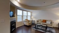 This well-appointed single-floor residence showcases a blend of elegant architectural details and comfortable living spaces. The standout feature of the living area is its striking curved ceiling design, which creates a sense of fluid, open movement throughout the room and adds a unique architectural character rarely found in residential spaces.

Rich, dark hardwood floors run throughout the living area, providing a warm and sophisticated foundation that complements the neutral color palette of the walls and furnishings. A bank of large windows dressed with horizontal blinds floods the space with abundant natural light, creating a bright and inviting atmosphere while offering views to the outside.

The living room is thoughtfully arranged with a neutral-toned sofa accented with warm orange and patterned throw pillows, paired with a matching armchair to create a cozy conversational setting. A glass-top coffee table and patterned area rug anchor the seating arrangement, adding layers of texture and visual interest. Recessed lighting in the curved ceiling tray supplements the natural light, ensuring the space is well-lit at any time of day. The overall ambiance is warm, modern, and welcoming, making it an ideal space for both relaxing and entertaining.