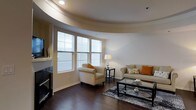 This well-appointed single-floor residence showcases a blend of elegant architectural details and comfortable living spaces. The standout feature of the living area is its striking curved ceiling design, which creates a sense of fluid, open movement throughout the room and adds a unique architectural character rarely found in residential spaces.

Rich, dark hardwood floors run throughout the living area, providing a warm and sophisticated foundation that complements the neutral color palette of the walls and furnishings. A bank of large windows dressed with horizontal blinds floods the space with abundant natural light, creating a bright and inviting atmosphere while offering views to the outside.

The living room is thoughtfully arranged with a neutral-toned sofa accented with warm orange and patterned throw pillows, paired with a matching armchair to create a cozy conversational setting. A glass-top coffee table and patterned area rug anchor the seating arrangement, adding layers of texture and visual interest. Recessed lighting in the curved ceiling tray supplements the natural light, ensuring the space is well-lit at any time of day. The overall ambiance is warm, modern, and welcoming, making it an ideal space for both relaxing and entertaining.