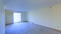 This single-floor living space presents a clean, open layout with a neutral aesthetic well-suited for a variety of furniture arrangements. The room is finished with wall-to-wall carpet in a soft, light tone that adds warmth underfoot and contributes to the overall understated color palette of white and grey throughout.

A sliding glass door along one wall serves as both a natural light source and a point of access to an outdoor balcony. Vertical blinds are installed, allowing for flexible light control and privacy. The view through the glass suggests a lush, tree-lined outdoor setting, bringing a sense of connection to the outdoors.

Heating is provided by a baseboard unit positioned along the lower portion of the far wall, a practical and unobtrusive feature common in residential spaces. The walls are smooth and painted in a crisp white, offering a blank canvas for personalization. Overall, the space conveys a functional, well-maintained environment with ample room to accommodate a comfortable living area arrangement.