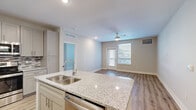 This thoughtfully designed single-floor residence in North Kansas City offers three bedrooms and two bathrooms within a comfortable and efficiently organized floor plan. The open-concept kitchen and living area serves as the heart of the home, anchored by a granite-topped island with a double sink and ample storage. The kitchen is fully equipped with gray shaker-style cabinetry, stainless steel appliances including a refrigerator, range, and microwave, and a mosaic tile backsplash that adds a stylish accent. Light wood-look flooring runs throughout the main living areas, complementing the neutral-toned walls and recessed lighting. The living room benefits from natural light streaming through a large window and a sliding door, with a ceiling fan adding comfort. The bedrooms are carpeted and bright, each featuring windows with blinds. The primary bedroom offers a generous footprint with its own en-suite bathroom, which includes a dual-sink granite vanity with gray cabinetry and wood-look flooring. The second bathroom features a walk-in shower with built-in shelving. Two closets and a hallway complete the layout, providing practical storage throughout this clean, modern residence.