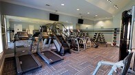 This fitness center is designed to provide a comprehensive workout experience within a single-floor layout. The space is outfitted with a robust selection of cardio equipment, including treadmills, elliptical trainers, and stair climbers, arranged in organized rows to maximize usability and flow. A dedicated free weights section lines one wall, offering a range of dumbbells suited to various fitness levels. Floor-to-ceiling mirrors span the perimeter of the room, enhancing the sense of openness and allowing users to monitor their form during exercise. Recessed lighting illuminates the space evenly throughout, complemented by wall-mounted flat-screen televisions positioned for easy viewing from the cardio equipment. The flooring features a durable, patterned carpet designed to withstand heavy foot traffic while providing comfort underfoot. The walls are finished in a cool, muted blue-gray tone that contributes to a calm and focused atmosphere. Natural light filters in through windows along the walls, further brightening the interior. The overall ambiance is clean, modern, and functional, making this fitness center a well-appointed facility suited for a full range of workout routines.