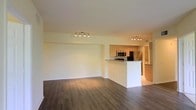 This single-floor space showcases an open-concept layout that connects the living area and kitchen in a seamless, functional design. The living area is generously proportioned, featuring rich, dark hardwood-style flooring that runs throughout and creates a warm, grounded ambiance. Crisp white walls and a smooth ceiling contribute to the bright, airy feel of the space.

The kitchen is positioned at the heart of the unit, anchored by a peninsula-style counter that serves as both a workspace and a natural divider between the cooking and living areas. Warm-toned cabinetry provides ample storage, while stainless steel appliances — including a refrigerator and microwave — lend a modern, polished character. Track lighting fixtures are installed in both the kitchen and the adjacent living area, offering focused, adjustable illumination throughout.

The transition between the kitchen and the broader living space is smooth and intuitive, making the layout ideal for everyday living and entertaining. The consistent flooring and neutral color palette tie the entire space together, creating a cohesive and well-maintained environment. Overall, the unit presents a clean, contemporary aesthetic with practical design choices throughout.