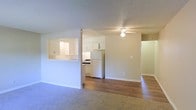 This single-floor residence features an open and functional layout designed to maximize the sense of space. The living area is finished with wall-to-wall carpeting and includes a ceiling fan, providing comfort and airflow. A pass-through window connects the living space to the kitchen, enhancing the open feel while maintaining a defined separation between areas.

The kitchen is outfitted with white cabinetry offering ample storage, a white refrigerator, and a range, all set against a clean, neutral color palette. The pass-through counter adds convenience for serving and casual interaction between rooms.

Adjacent to the carpeted living area, a hallway leads to additional rooms, where hardwood-style flooring continues, adding a cohesive visual flow throughout the unit. The walls are painted in soft, neutral tones, complementing the light-colored cabinetry and white appliances.

The textured ceilings are consistent throughout the space, adding subtle dimension overhead. Overall, the layout is straightforward and practical, with a bright, clean aesthetic that creates a welcoming and livable environment.