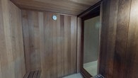 This single-level sauna space is thoughtfully constructed with floor-to-ceiling vertical wood paneling that extends across all walls and the ceiling, creating a cohesive and warm aesthetic throughout. The rich, natural tones of the wood lend the space a classic, cabin-like ambiance that is both functional and inviting.

The main sauna room is equipped with tiered wooden benches, allowing users to choose their preferred heat level by adjusting their seating position. A traditional stone heater sits in the corner of the room, framed by a metal safety guard and loaded with heat-retaining rocks — the hallmark of an authentic Finnish-style sauna experience. A wall-mounted thermometer is prominently positioned, allowing for precise temperature monitoring during use. Soft, warm lighting is provided by a single wall-mounted fixture, casting a gentle glow across the wood-paneled surfaces.

A full-length glass door with a dark wood frame separates the sauna from an adjacent shower area, which features clean, light-colored tile walls and flooring. This seamless connection between the sauna and shower facilitates a convenient and complete heat therapy routine. The mosaic tile flooring at the sauna's entrance adds a subtle design detail while providing a practical, water-resistant transition surface. Overall, the space is designed for comfort, relaxation, and ease of use.