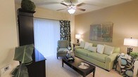 This single-floor residence offers a thoughtfully designed living environment centered around a welcoming and well-lit living room. The space is anchored by a large sliding glass door dressed with vertical blinds and decorative patterned curtains, allowing generous natural light to fill the room while providing privacy and style. A classic five-blade ceiling fan with a light fixture overhead ensures year-round comfort and adds a timeless touch to the layout. The living room is finished with plush wall-to-wall carpeting underfoot and warm, neutral-toned walls that create a calm and inviting ambiance. Furnishings include a sage green sofa accented with coordinating pillows, a matching upholstered armchair, and a sleek dark-wood coffee table that grounds the seating arrangement. A side table with a lamp provides warm accent lighting, complementing the overall cozy atmosphere. A dark wood storage unit or entertainment console anchors one side of the room, adding functional storage to the space. The combination of soft tones, natural light, and carefully chosen furnishings gives this residence a comfortable, move-in-ready feel that balances practicality with a welcoming residential charm.