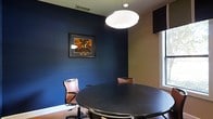 This single-floor space presents a refined and professional atmosphere, highlighted by a well-appointed meeting or conference room. The room's most striking feature is its deep navy blue accent walls, which create a bold, focused environment suited for professional gatherings or private meetings. A large, round dark-topped table serves as the centerpiece, surrounded by ergonomic brown leather chairs that add warmth and contrast against the cool wall tones. Overhead, a statement globe pendant light provides soft, even illumination, complementing the room's overall ambiance. Natural light filters in through a window fitted with a dark valance, offering a balanced blend of artificial and natural lighting. The warm hardwood flooring grounds the space, adding texture and continuity. A framed piece of artwork with rich amber and brown tones adorns the navy wall, providing a curated visual focal point. The overall design strikes a balance between formality and comfort, making it well-suited for focused discussions, client meetings, or collaborative sessions. The cohesive color palette and deliberate furnishing choices reflect a thoughtfully designed professional environment.