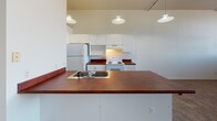 Located at 337 N Huron St in downtown Toledo, Ohio, this single-floor unit showcases an open, loft-inspired layout that blends functional design with urban character. The living area features neutral wall-to-wall carpeting and generously proportioned open space, giving the interior a roomy, flexible feel suitable for various configurations.

The kitchen is a notable feature, highlighted by rich, deep-toned countertops that extend across both the peninsula and perimeter surfaces, creating a striking contrast against the white cabinetry and white appliances, which include a refrigerator and electric range. A series of pendant lights hang from the ceiling above the peninsula, adding warmth and a contemporary touch. The open-plan connection between the kitchen and living area promotes an easy flow throughout the space.

Industrial elements are woven throughout the unit, including exposed ductwork and piping along the ceilings, which lend a distinctly urban aesthetic. Track lighting in the main areas supplements natural light, which enters through a bank of windows offering views of the surrounding cityscape.

The bathroom is clean and practical, featuring a vanity with ample cabinet storage, a large mirror, and a tub-shower combination. Overall, the unit presents a straightforward, urban living environment in the heart of Toledo's downtown corridor.