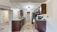 This single-floor unit in Holly, Michigan offers a well-designed layout across two bedrooms and two bathrooms. The kitchen is a standout feature, equipped with rich dark-stained cabinetry, granite countertops, and a full suite of stainless steel appliances including a refrigerator, range, and microwave. A peninsula with an undermount sink and additional granite surface extends into the main living area, creating a seamless connection between the kitchen and the open living space. Light wood-style flooring runs throughout the kitchen and main areas, while plush carpet adds warmth to the living room and bedrooms.

The living room is generously proportioned, featuring a ceiling fan and a sliding glass door that opens to a private balcony with pleasant outdoor views. The primary bedroom includes a walk-in closet and an en-suite bathroom, while the second bedroom offers its own closet and easy access to the second full bath. Additional practical features include a utility room with built-in shelving, a water heater, and an electrical panel. Neutral gray tones throughout create a clean, modern aesthetic, and the thoughtfully designed floor plan ensures comfortable, functional living throughout the unit.