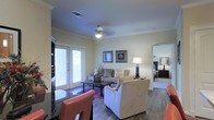 This single-story space showcases a well-designed open-concept layout that seamlessly connects the dining and living areas. The living room is anchored by hardwood-style flooring and features elegant crown molding throughout, adding a refined architectural touch. A ceiling fan with integrated lighting serves as a functional centerpiece overhead, while an HVAC vent ensures year-round comfort.

The living area is bright and inviting, benefiting from natural light streaming through a set of French doors that lead to an outdoor space. The room comfortably accommodates a sofa, armchair, and coffee table arrangement, creating a welcoming seating area. Two framed artworks adorn the walls, complemented by table lamps that add warm ambient lighting.

In the foreground, a dining area with dark seating is partially visible, suggesting a practical transition between the kitchen and living spaces. An arched doorway leads to an adjacent bedroom, which is partially visible and features carpeted flooring, offering a contrast to the main living area's hard surface floors. The overall ambiance is calm and functional, with neutral wall tones enhancing the sense of openness.