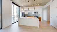 Located in the heart of downtown Salt Lake City at 131 S 300 W, this contemporary 3-bedroom, 2-bathroom residence showcases an industrial-modern aesthetic defined by exposed concrete ceilings, light hardwood flooring, and crisp white walls. The open-concept living, dining, and kitchen area serves as the centerpiece of the home, anchored by a large waterfall-edge island and a sleek kitchen outfitted with stainless steel appliances, warm wood cabinetry, a glass subway tile backsplash, and gold pendant lighting. Floor-to-ceiling windows flood the space with natural light and frame sweeping views of the Salt Lake City skyline and surrounding cityscape. The bedrooms feature plush carpet, exposed concrete columns, and large windows that continue the urban aesthetic, with ample closet storage available across four closets. The primary bedroom offers generous proportions with its own access to closets. The two bathrooms are elegantly appointed with slate-toned subway tile, glass-enclosed showers, and light wood vanities. One bathroom includes both a soaking tub and a separate walk-in shower. A clean hallway with modern panel doors connects the private quarters to the main living area, completing this well-designed urban residence.