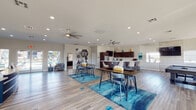 This single-story community clubhouse is designed with a modern, open-concept layout that encourages social interaction and comfortable everyday living. The main gathering area is expansive and light-filled, featuring wood-style plank flooring, recessed lighting, and dual ceiling fans that keep the atmosphere fresh and inviting. Vibrant teal area rugs anchor clusters of high-top tables and bar-stool seating, creating versatile spaces for casual dining or socializing.

A standout curved kitchen bar serves as the centerpiece of the space, finished in rich dark wood cabinetry with a sleek white countertop and complemented by modern swivel bar stools. The kitchen is fully equipped with stainless steel appliances, ample upper cabinetry, and colorful decorative accents. A dedicated beverage and snack station with matching dark wood cabinetry provides added convenience.

A business center nook offers a built-in desk workstation beneath wide windows, providing natural light and outdoor views — ideal for residents working remotely. The space also includes a billiards table positioned beneath large windows, adding a recreational element to the clubhouse.

The reception area features a textured stone accent wall, a branded front desk, and teal color-blocked walls that lend a polished, professional feel. Large sliding glass doors throughout the space open to outdoor views, ensuring a bright and connected atmosphere year-round.