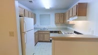 This single-floor space centers around a well-organized kitchen that offers a practical and efficient layout. The kitchen features light wood cabinetry that lines the walls, providing generous storage both above and below the countertops. The neutral-toned countertops extend across an L-shaped configuration, offering ample workspace for meal preparation. A gas range cooktop is conveniently positioned along one counter, complemented by an overhead ventilation hood, while a full-size white refrigerator anchors the opposite wall. A stainless steel sink is situated beneath a window fitted with horizontal blinds, allowing natural light to filter into the space. A peninsula counter extends outward, adding additional prep or serving space and subtly defining the kitchen area. The light-tiled flooring is easy to maintain and contributes to the clean, airy feel of the room. A textured ceiling adds a classic touch overhead. The overall ambiance is straightforward and functional, with a neutral color palette of soft blues and warm wood tones creating a calm, approachable environment suited to everyday use.