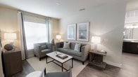 This single-floor residence offers a thoughtfully designed living space characterized by a clean, contemporary aesthetic and a warm, neutral color palette. The living area serves as the heart of the home, featuring wood-look flooring that adds warmth and durability underfoot, complemented by a soft area rug that defines the seating arrangement. Large windows dressed with white blinds and curtain panels flood the room with natural light, creating a bright and welcoming ambiance throughout the day. The space is furnished with a comfortable upholstered sofa accented by striped throw pillows, a coordinating armchair, and a sleek coffee table with a metal base and marble-top surface. Tasteful framed artwork adorns the walls, adding visual interest without overwhelming the serene atmosphere. Recessed ceiling vents ensure efficient climate control throughout the space. The overall design strikes a balance between modern minimalism and comfortable livability, making it an inviting environment for both relaxing and entertaining. Neutral walls provide a versatile backdrop that allows the furnishings and natural light to take center stage, resulting in a cohesive and polished interior throughout.