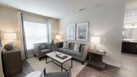 This single-floor residence offers a thoughtfully designed living space characterized by a clean, contemporary aesthetic and a warm, neutral color palette. The living area serves as the heart of the home, featuring wood-look flooring that adds warmth and durability underfoot, complemented by a soft area rug that defines the seating arrangement. Large windows dressed with white blinds and curtain panels flood the room with natural light, creating a bright and welcoming ambiance throughout the day. The space is furnished with a comfortable upholstered sofa accented by striped throw pillows, a coordinating armchair, and a sleek coffee table with a metal base and marble-top surface. Tasteful framed artwork adorns the walls, adding visual interest without overwhelming the serene atmosphere. Recessed ceiling vents ensure efficient climate control throughout the space. The overall design strikes a balance between modern minimalism and comfortable livability, making it an inviting environment for both relaxing and entertaining. Neutral walls provide a versatile backdrop that allows the furnishings and natural light to take center stage, resulting in a cohesive and polished interior throughout.