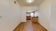 This single-floor space features an open-concept layout that seamlessly connects the living area to the kitchen, creating a cohesive and functional floor plan. The living area is bright and airy, with tall ceilings that enhance the sense of space, neutral white walls, and warm hardwood-style flooring that extends throughout. A ceiling-mounted light fixture provides overhead illumination, while a ceiling vent ensures climate control comfort.

The kitchen is tucked into a defined alcove and is well-equipped with medium-toned wooden cabinetry offering ample storage above and below the counters. A white range and under-cabinet range hood anchor the cooking area, complemented by a dark-countered sink with a window above. Dark countertops provide contrast against the lighter cabinetry. A peninsula with a matching dark countertop and wooden base separates the kitchen from the living space, offering additional prep area or casual seating. The overall ambiance is clean, practical, and welcoming, with a warm color palette created by the interplay of natural wood tones, neutral walls, and consistent flooring.