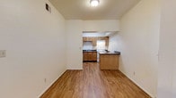 This single-floor space features an open-concept layout that seamlessly connects the living area to the kitchen, creating a cohesive and functional floor plan. The living area is bright and airy, with tall ceilings that enhance the sense of space, neutral white walls, and warm hardwood-style flooring that extends throughout. A ceiling-mounted light fixture provides overhead illumination, while a ceiling vent ensures climate control comfort.

The kitchen is tucked into a defined alcove and is well-equipped with medium-toned wooden cabinetry offering ample storage above and below the counters. A white range and under-cabinet range hood anchor the cooking area, complemented by a dark-countered sink with a window above. Dark countertops provide contrast against the lighter cabinetry. A peninsula with a matching dark countertop and wooden base separates the kitchen from the living space, offering additional prep area or casual seating. The overall ambiance is clean, practical, and welcoming, with a warm color palette created by the interplay of natural wood tones, neutral walls, and consistent flooring.