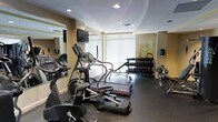This fitness center is thoughtfully designed to accommodate a full range of exercise needs within a clean, functional layout. The space features a rubberized dark flooring that provides both durability and comfort underfoot, making it well-suited for high-impact workouts. A selection of cardio equipment takes center stage, including elliptical trainers and treadmills from Star Trac, a reputable fitness brand known for commercial-grade performance. For strength training, a multi-tiered dumbbell rack lines one side of the room, offering a comprehensive range of free weights to support various fitness levels. A wall-mounted flat-screen television is positioned for easy viewing during cardio sessions, enhancing the overall workout experience. Large frosted windows along one wall allow diffused natural light to fill the space while maintaining privacy. Recessed ceiling lighting supplements the natural light, ensuring the room remains bright and well-illuminated at all times. A full-length mirror on one wall visually expands the space and allows for proper form checks during exercise. The neutral warm-toned walls and white ceiling contribute to a clean, professional ambiance consistent with a well-maintained fitness facility.