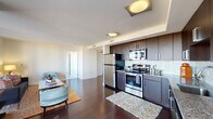 This single-floor residence showcases a thoughtfully designed open-concept layout with rich dark hardwood flooring throughout the main living areas. The living room is anchored by expansive floor-to-ceiling windows that frame sweeping elevated views of the surrounding landscape, flooding the space with abundant natural light. A comfortable seating arrangement with light blue accent chairs and a dark sofa centers the room around a marble-topped coffee table, complemented by a warm area rug.

Adjacent to the living area, a dedicated dining space accommodates an intimate round table and chairs, benefiting from the same dramatic window views. The kitchen features dark espresso cabinetry, granite countertops, a white subway tile backsplash, and a full suite of stainless steel appliances, including a refrigerator, range, and built-in microwave.

The bedroom offers a calm, neutral retreat with carpeted flooring and is accompanied by a large built-in closet system with dark wood shelving and hanging rods providing generous storage. Two bathrooms complete the unit — one with a tub-shower combination and granite vanity, and another with a streamlined vanity and tile flooring — both finished with dark wood cabinetry and modern fixtures.