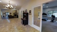 This single-story space showcases a well-connected, open floor plan with consistent light beige ceramic tile flooring throughout. The main living area is visible to the left, anchored by a stacked-stone fireplace that serves as a warm, prominent focal point. Recessed lighting is installed throughout the ceiling, providing even, ambient illumination across the space.

A built-in dark wood hutch and compact desk area occupy a central wall, offering a practical workspace or organizational station. Small decorative accents, including framed artwork and wall hangings, add a personalized touch to the neutral-toned walls.

A standout architectural feature is the large glass-panel French door, framed in white, which opens into a dedicated exercise or fitness room. This transparent partition allows natural sightlines between spaces while maintaining a degree of separation. The fitness room, visible through the glass, appears to house exercise equipment and benefits from its own recessed lighting.

A louvered door nearby suggests additional storage or utility access. The overall ambiance is practical and comfortable, with a clean, neutral palette that allows for flexible use throughout the single-level layout.