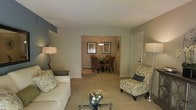 This single-floor residence offers a thoughtfully arranged layout that balances comfort and functionality. The living area serves as the heart of the home, featuring plush carpeted flooring in a warm neutral tone that extends throughout the space. A glass-topped coffee table anchors the seating area, complemented by a classic white sofa adorned with decorative pillows and an elegant accent chair upholstered in a floral pattern. Soft ambient lighting is provided by stylish floor and table lamps, creating a welcoming atmosphere. The living area opens directly into a cozy dining space, visible through a wide passage that enhances the sense of connectivity between rooms. The dining area is appointed with a round dark-wood table surrounded by cushioned chairs, overhead pendant lighting, and framed wall art that adds a touch of sophistication. The walls throughout are painted in soothing greige tones with subtle accent detailing, contributing to a calm and cohesive aesthetic. A white entry door is conveniently positioned near the living area, providing easy access. The overall ambiance is warm, inviting, and move-in ready, making it an ideal space for both relaxing and entertaining.