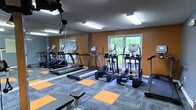 This fitness center offers a dedicated space for cardiovascular and general exercise, thoughtfully designed with both functionality and comfort in mind. The room is finished with bold orange accent walls paired with gray trim, creating an energizing atmosphere that complements the active environment. A patterned carpet floor featuring alternating gray and orange tiles adds to the vibrant aesthetic while providing a practical, durable surface underfoot.

The space is outfitted with a selection of cardio equipment, including multiple treadmills and elliptical trainers from the TRUE Fitness brand, arranged to maximize floor space and allow for comfortable use between machines. A full-length mirror spans one wall, visually expanding the room and allowing users to monitor their form during workouts.

Natural light filters in through multiple windows along the exterior wall, offering pleasant views of the surrounding greenery outside. Overhead fluorescent lighting ensures the space remains bright and well-lit at all times. The room is also equipped with a mounted television, adding an entertainment option for users during their workouts. The overall ambiance is clean, modern, and motivating, making it a practical and inviting facility for daily fitness routines.
