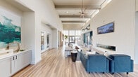 Located at 13611 Perry Rd in Houston, TX 77070, this single-story community clubhouse delivers an impressive blend of modern architecture and thoughtfully curated interior design. The space is defined by its dramatic soaring ceilings with exposed beam detailing and an abundance of natural light streaming through expansive floor-to-ceiling windows and glass doors, offering views to the outdoor surroundings.

Wide-plank wood-look flooring runs continuously throughout, anchoring the open-concept layout that flows seamlessly from one area to the next. The main lounge area features plush seating arrangements, including deep blue velvet sofas and neutral accent chairs, complemented by a sleek linear fireplace set against a bold teal feature wall. Curated artwork and built-in display shelving add visual interest at elevation.

A dedicated dining and social area is equipped with a large wood communal table, pendant lighting, and a stylish dark-toned backdrop, creating an inviting atmosphere for gatherings. A cozy built-in banquette nook, adorned with a patterned blue wallcovering and warm wood surround, provides an intimate seating option. The entry foyer greets visitors with tall vertical artwork, live plants, and a welcoming seating cluster, setting a refined tone from arrival.
