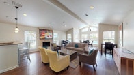 This single-floor community space is designed with an open-concept layout that seamlessly connects the living, dining, and kitchen areas into one expansive, welcoming environment. The soaring vaulted ceilings lend a sense of grandeur, while rich hardwood flooring runs throughout, adding warmth and cohesion to the interior.

The main living area is anchored by a brick fireplace framed by a vibrant large-scale artwork, creating a focal point that draws the eye. Comfortable seating — including a taupe sofa accented with teal throw pillows and a pair of soft yellow armchairs — is arranged around a sleek metal coffee table, encouraging relaxed conversation. Recessed lighting and an abundance of natural light from multiple windows keep the space bright and airy.

Adjacent dining zones feature dark wood tables and chairs, with large arched windows offering picturesque views of an outdoor pool and surrounding greenery. The kitchen is well-appointed with warm cherry wood cabinetry, granite-look countertops, black appliances, and a breakfast bar with seating for four, lit by pendant lighting overhead. A secondary kitchen area provides additional prep and sink space with tiled flooring. Throughout, the neutral color palette is enlivened by pops of teal and orange in the décor, lending the space a modern, inviting character.