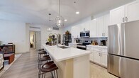 Located in Gilbert, AZ, this thoughtfully designed 2-bedroom, 2-bathroom single-story home offers a well-appointed open-concept layout. The kitchen is a standout feature, showcasing crisp white shaker cabinetry, a decorative tile backsplash, stainless steel appliances, and a generous island with bar seating for four. Geometric pendant lights add a modern touch overhead, while light wood-style flooring flows seamlessly throughout the space. The adjacent living room is bright and welcoming, featuring French doors that invite natural light and provide access to an outdoor area. The bedrooms feature distinctive sage green accent walls, creating a calming atmosphere. The primary bedroom includes a sliding barn door leading to a closet, while both rooms benefit from natural light through large windows. Each bathroom is finished with white cabinetry and light countertops; one features a tub-shower combination, while the other includes a walk-in glass-enclosed shower with large-format tile. The dedicated laundry room is well-organized with stacked washer and dryer units and open shelving for storage. This home combines modern design with practical functionality in a comfortable, single-level floor plan.