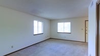 This single-floor space presents a straightforward and functional layout. The main room visible is spacious and open, featuring neutral-toned walls that create a clean, versatile backdrop suitable for a variety of uses. The room is finished with wall-to-wall carpeting in a light gray tone, adding a degree of warmth and comfort underfoot throughout the space.

Two windows are positioned along adjacent walls, allowing natural light to filter into the room from multiple directions. The windows are fitted with horizontal blinds, providing practical light control. The placement of these windows contributes to a bright atmosphere, even on overcast days, as light reflects evenly off the pale walls.

The ceiling features a textured finish, a common and durable detail that adds subtle visual depth. Dark wood baseboards line the perimeter of the room, providing a mild contrast to the light walls and carpet, and adding a modest degree of architectural definition.

Electrical outlets are positioned at standard intervals along the walls, supporting flexible furniture arrangement. Overall, the space maintains a simple, neutral aesthetic that serves as a functional and adaptable environment.