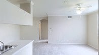 This single-floor residential space offers a clean, open layout defined by a cohesive neutral color palette throughout. The living area is generously proportioned, with plush wall-to-wall carpeting underfoot and smooth ceilings accented by classic crown molding that adds a refined architectural touch. A ceiling fan is installed overhead, contributing to both comfort and airflow. One of the standout design elements is the graceful arched doorway that connects the main living area to the adjacent hallway, lending a subtle elegance to the interior architecture. The walls are finished in a warm beige tone that creates a calm, inviting ambiance. A raised dividing counter or half-wall separates the living space from what appears to be a kitchen or dining area, providing an open yet defined floor plan. HVAC vents are integrated into the walls and ceiling, and a thermostat is conveniently positioned along one wall. The hallway beyond the arched entry leads to additional rooms, suggesting a well-organized layout with clear separation between the living and private areas. Overall, the space presents a functional and welcoming environment with timeless design details suited for comfortable everyday living.