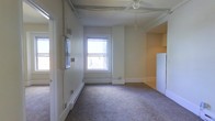 This single-floor space offers a functional and straightforward layout designed for practical everyday living. The main living area is finished with wall-to-wall carpeting in a neutral tone, creating a comfortable and cohesive feel underfoot. A ceiling fan with a light fixture is centrally mounted, providing both air circulation and ambient lighting throughout the room. Natural light enters through a double-hung window, brightening the space and offering a view to the exterior. Baseboard heating units line the perimeter walls, ensuring efficient warmth during cooler months. Exposed conduit and track lighting along the upper walls add an industrial touch to the otherwise clean, light-painted interior. Adjacent to the main living area, a compact kitchen nook is partially defined by upper cabinetry in a warm wood finish and includes a full-size white refrigerator, providing essential storage and appliance space. White baseboard trim throughout lends a crisp, finished appearance to the walls. The overall ambiance is modest and utilitarian, making the space well-suited for those seeking a no-frills, move-in-ready environment with all the core elements in place.