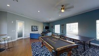 This single-floor recreational space is designed with both leisure and social gathering in mind. The room's most prominent feature is a branded "Reserve" pool table, centered on a bold blue geometric area rug over rich hardwood flooring, giving the space a polished yet inviting character. Adjacent to the pool table sits a ping pong table, making this a versatile game room suited for a variety of activities.

The lounge area is furnished with contemporary blue sofas and chairs, complemented by multiple wall-mounted flat-screen televisions, creating an ideal setting for watching sports or casual entertainment. Recessed ceiling lighting and a ceiling fan with a warm-toned fixture provide both functional illumination and a comfortable atmosphere throughout the space.

The walls are painted in a crisp light gray with a deep teal accent wall that adds a modern, dynamic contrast to the room's design. Crown molding along the ceiling adds a refined architectural touch. Natural light filters in through multiple windows fitted with horizontal blinds, offering views of the surrounding exterior while maintaining privacy. The overall ambiance is energetic yet comfortable, reflecting a community-oriented space designed to encourage interaction and recreation.