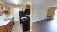 This single-floor residential unit offers a practical and well-organized layout designed for comfortable everyday living. The kitchen is equipped with a full suite of black appliances, including a refrigerator, electric coil range, over-the-range microwave, and dishwasher, all complemented by warm wood-toned upper cabinetry that provides ample storage. The kitchen's light yellow walls create a bright, welcoming atmosphere, while the hardwood-style laminate flooring extends seamlessly throughout the space, adding warmth and visual continuity. A baseboard heating unit lines the wall of the adjoining open area, which flows naturally from the kitchen and offers flexible use as a dining or living space. The layout is efficient, making excellent use of available square footage. Recessed and overhead lighting ensures the interior remains well-lit. A doorway visible from the kitchen suggests additional rooms beyond, contributing to the overall functionality of the floor plan. Clean white trim and neutral wall tones throughout the space create a cohesive, move-in-ready aesthetic suitable for a variety of furnishing styles.