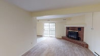 This single-floor residence offers a well-organized layout with distinct living, sleeping, and functional zones. The living area is the centerpiece of the home, featuring a classic brick fireplace with a white mantle and brass-accented screen, soft neutral carpet underfoot, and a structural beam that subtly defines the space. A large sliding glass door fills the room with natural light and opens to a private balcony, offering outdoor views. Baseboard heating runs along the perimeter walls throughout the unit.

The kitchen is equipped with warm wood cabinetry, a white electric range, a refrigerator, and a countertop sink. Light tile flooring and efficient overhead lighting give the kitchen a clean, functional character.

The bedrooms are carpeted and finished in soft cream-toned walls, offering a calm and neutral atmosphere. Each room benefits from natural light through windows, with one bedroom featuring a large multi-pane window with a baseboard heater beneath it and pleasant outdoor views.

The bathroom includes a tiled tub-and-shower combination with white tile surround, a basketweave tile floor, a vanity with wood cabinetry, and a wall-mounted mirror with overhead lighting. The space is clean and practical in design throughout.