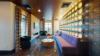 This boldly designed lounge space makes an immediate impression through its fearless use of color, texture, and artistic detail. The most commanding feature is a floor-to-ceiling accent wall lined with rows of illuminated disc sconces set against a deep teal background, casting a warm, golden glow that defines the room's vibrant character. Beneath, a plush tufted sofa in rich purple provides generous seating, complemented by rounded wood coffee tables and teal accent chairs arranged throughout the space.

The flooring is a polished terrazzo in deep, speckled tones that reflects light beautifully and adds to the retro-contemporary aesthetic. Overhead, a warm metallic ceiling amplifies the ambient lighting, creating a cohesive, moody atmosphere. Plaid drapery panels frame large windows that invite abundant natural light and frame city skyline views.

A wall-mounted flat-screen television and a sleek fireplace add functional elegance to one side of the lounge. Glass double doors connect the interior to an outdoor terrace, where additional seating takes advantage of open-air city views. An adjacent area, visible through the glass-paneled entry, features additional furnishings and decorative wall graphics. The overall ambiance is energetic yet sophisticated, blending artistic flair with comfortable, functional design.