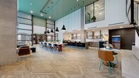 Located at 4015 Craft St in Charlotte, NC, this single-floor community amenity space is a masterclass in contemporary design. The centerpiece is a dramatic, double-height open-concept great room with soaring ceilings accented by teal architectural detailing and floor-to-ceiling windows that flood the interior with natural light and offer views of the outdoor pool courtyard. Herringbone-patterned hardwood flooring runs throughout the main gathering area, lending warmth and texture underfoot.

A large communal dining table anchors the social hub, complemented by bar-height seating along a sleek kitchen counter. The full kitchen is outfitted with weathered-wood cabinetry, open iron-framed shelving, a geometric black-and-white tile floor, concrete countertops, and warm Edison-bulb pendants—combining industrial and organic elements with refined style.

A dedicated lounge area offers generous sectional seating in leather and fabric, positioned beneath a sculptural cluster of globe pendant lights and oriented toward oversized windows. A media lounge with dark, paneled walls and a built-in entertainment console with large-screen displays provides a cozy retreat. A game room featuring a shuffleboard table and bold geometric tile flooring rounds out the entertainment offerings. Throughout, the design balances bold contrasts with an inviting, sophisticated ambiance.