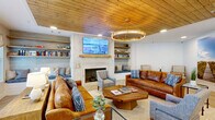 Located at 49 N Assembly Dr in Wilmington, NC, this single-floor communal space offers a generously sized, cohesive layout that blends coastal charm with refined interior design. The living room is a standout centerpiece, featuring a striking tongue-and-groove wood ceiling, a statement ring chandelier, and a textured stone fireplace flanked by open wooden shelving and a built-in window bench. Rich cognac leather sofas, a solid wood coffee table, and layered soft furnishings create a warm, inviting atmosphere. A dedicated reading nook with shiplap walls, floating shelves stocked with books, and upholstered accent chairs with ottomans provides a quiet retreat within the larger space. An adjacent lounge area opens to the outdoors through large floor-to-ceiling glass doors and windows, flooding the interior with natural light and offering views of the surrounding community. Light wood herringbone flooring transitions seamlessly throughout. The dining room features a rustic round table set within a cozy banquette with wood-paneled backing, topped by an elegant cage chandelier. Coastal artwork and a whitewashed display cabinet complete the refined, beach-inspired aesthetic. The overall ambiance is welcoming, well-appointed, and thoughtfully curated.