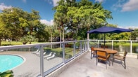 Located at 1 South Pine Island Road in Plantation, Florida, this outdoor terrace offers a refined and inviting al fresco experience surrounded by the lush tropical landscape characteristic of South Florida. The terrace is enclosed by a sleek glass-panel railing with a clean metal frame, providing unobstructed sightlines to the verdant grounds below while maintaining an open, airy feel.

The space overlooks a pool area furnished with lounge chairs, offering a relaxed resort-like atmosphere. A round outdoor dining table paired with tan-and-dark-framed chairs is positioned on the terrace, shaded by a navy blue market umbrella — perfect for casual outdoor dining or socializing. The concrete deck underfoot is well-maintained and complements the modern aesthetic of the railing system.

Mature trees, including sweeping palms and broad canopy trees, frame the backdrop, creating a natural privacy screen and adding to the lush, park-like setting. The terrace benefits from abundant natural light and open sky views, with the greenery providing welcome shade and a tranquil ambiance.

Overall, this terrace delivers a seamlessly connected indoor-outdoor experience, combining practical amenities with a serene, tropical outdoor environment in the heart of Plantation, FL.