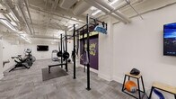 This fitness center is a thoughtfully designed, single-floor facility built to support a wide range of workout routines. The space is organized into distinct functional zones, allowing users to move seamlessly between cardio, strength training, and functional fitness activities.

The cardio area is outfitted with a treadmill, elliptical trainer, rowing machine, and stationary bikes, including an air bike. A wall-mounted flat-screen TV is positioned for easy viewing during workouts. Overhead, exposed ductwork and industrial-style fluorescent lighting give the space a clean, utilitarian aesthetic with white walls that keep the environment feeling open and bright.

The strength and functional training zone features a multi-purpose power rack equipped with barbells, weight plates, gymnastic rings, suspension straps, and battle rope attachments. A flat utility bench accompanies the rack, and a dumbbell rack stocked with a range of free weights rounds out the strength equipment offerings.

Bold accent walls in deep purple and warm amber add a motivating, energetic atmosphere to the space, complemented by a motivational mural that reinforces a high-performance mindset. The flooring throughout consists of durable carpet tiles with rubber mat overlays in the lifting areas for added safety and stability.

The facility also includes a dedicated exit, a waste station, and sanitation supplies, ensuring a clean and functional environment for all users.