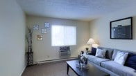 This single-floor residential space offers a comfortable and functional layout designed for everyday living. The living room serves as the heart of the home, featuring wall-to-wall carpeting that adds warmth underfoot. A large window fitted with horizontal blinds allows natural light to fill the room, creating a bright and airy atmosphere. A wall-mounted HVAC unit beneath the window provides convenient climate control, while a baseboard heater along the wall ensures warmth during cooler months. The textured ceiling adds a subtle architectural detail overhead. The room provides ample space for a generously sized sofa accented with decorative pillows, accompanied by a sleek dark coffee table that anchors the seating area. Soft ambient lighting from a table lamp adds to the inviting ambiance of the space. Decorative wall art arranged in an asymmetric cluster brings personality and charm to the neutral-toned walls. The overall color palette — featuring light beige walls and neutral carpeting — creates a calm, versatile backdrop that lends itself well to a variety of interior styles. This space combines practicality with a cozy residential feel, making it a comfortable place to relax and unwind.