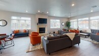 This single-story community clubhouse is thoughtfully designed to provide residents with a variety of functional and social spaces. The layout flows naturally from a welcoming lounge entry into several distinct areas, each finished with wood-look flooring, recessed lighting, and a neutral color palette that creates a bright, cohesive atmosphere throughout.

The main lounge area is generously sized, featuring vaulted ceilings, large windows that invite abundant natural light, and a fireplace framed by a wall-mounted television. Comfortable sectional seating, tufted upholstery, and accent chairs in warm orange tones give the space an inviting, residential feel. An adjacent seating nook provides a more intimate setting with a round dining table and a teal bench with coordinating accent pillows.

The kitchen is well-appointed with rich dark wood cabinetry, granite countertops, stainless steel appliances, and a wraparound breakfast bar with bar stool seating. Pendant lighting overhead adds a polished touch. A transition area with wood-look flooring connects this space to a service counter equipped with additional storage and small appliances.

The fitness center occupies a dedicated wing with rubber flooring, ceiling fans, and ample natural light from multiple windows. It is equipped with treadmills, ellipticals, a multi-function cable machine, a dumbbell rack, and a wall of mirrors, providing a comprehensive workout environment for all fitness levels.